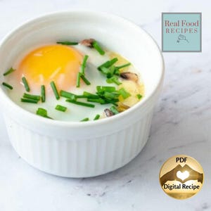 May include: A baked egg dish in a white ramekin. The egg yolk is bright yellow, surrounded by white egg whites, mushrooms, and green chives. The dish is presented on a white marble surface. Text in the image reads "Real Food Recipes" and "PDF Digital Recipe."