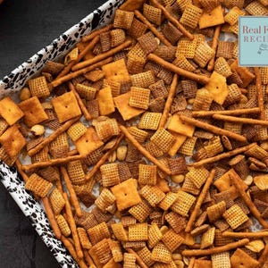 May include: A tray filled with a mix of snack foods, including square crackers, pretzel sticks, and peanuts. The snacks are various shades of brown and tan, with a few golden-colored crackers. The tray has a black and white speckled pattern. The text "Real Food Recipes" is visible.