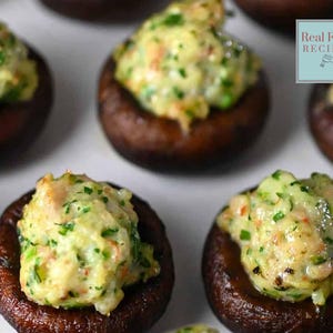 Puede incluir: Primer plano de tapas de champiñones rellenas de una mezcla cremosa y verde. Los champiñones están dispuestos sobre una superficie blanca. El texto "Real Food RECIPES" es visible en la esquina superior derecha de la imagen.