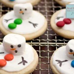 May include: Round sugar biscuits decorated with white icing to resemble snowmen. Each biscuit features a marshmallow head with a drawn face, orange nose, and two candy buttons in red, green, or blue. Black icing arms are drawn on the white surface.