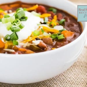 May include: A white bowl filled with chili, topped with sour cream, shredded cheddar cheese, and chopped green onions. The chili contains visible chunks of vegetables and beans. The image is a close-up shot, highlighting the textures and colors of the ingredients.