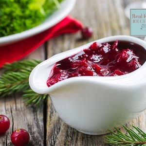 Puede incluir: Una salsera de cerámica blanca llena de salsa de arándanos rojos. La salsa es espesa y brillante, y la salsera está sobre una mesa de madera con ramas de pino verdes y arándanos rojos.