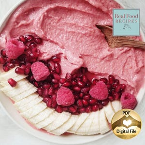 Könnte beinhalten: Eine rosa Smoothie-Bowl mit Himbeeren, Granatapfelkernen und Bananenscheiben. Die Schüssel trägt den Text "Real Food Recipes" und "PDF Digital File". Ein Holzlöffel liegt an der Seite. Das Essen ist auf einer weißen Oberfläche angerichtet.