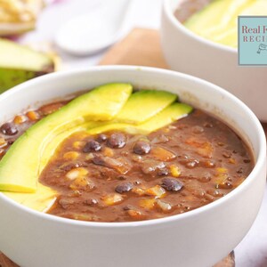 Puede incluir: Un tazón blanco lleno de sopa de frijoles y maíz de color marrón oscuro, cubierto con tres rodajas finas de aguacate verde brillante. El texto Real Food RECIPES es visible en un cuadrado azul claro en la esquina superior derecha.
