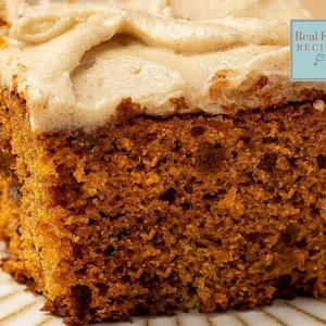 May include: A close-up of a slice of cake with light brown frosting. The cake is a rich, dark brown colour with a textured appearance. The frosting is thick and swirled on top. The image includes a small blue square with the text "Real Food RECIPES".