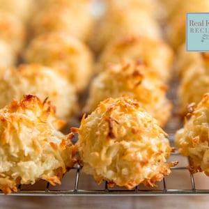 May include: A close-up of a wire rack with a batch of golden brown coconut macaroons. The macaroons are arranged in a single layer on the rack. The macaroons are slightly browned and have a fluffy texture.