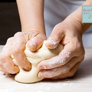 Puede incluir: Primer plano de manos amasando una bola de masa sobre una superficie blanca. La masa es blanca y lisa, y las manos están cubiertas de harina.