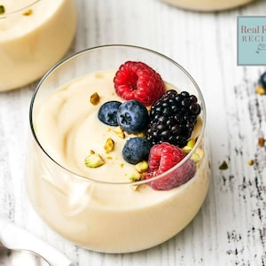 May include: A glass dessert dish filled with a creamy white pudding topped with fresh blueberries, raspberries, blackberries, and chopped pistachios. The dish is on a white wooden surface. "Real Food Recipes" is written in a blue box on the right side of the image.