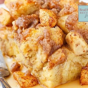 May include: A close-up of a casserole dish filled with a baked bread pudding with apples and a cinnamon-sugar topping. The dish is on a white plate with a fork and a small amount of sauce.