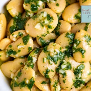 May include: A bowl of cooked butter beans tossed in a green herb and garlic oil dressing. The dish features large, pale, oval-shaped beans coated in a glossy sauce with visible flecks of green herbs and white garlic. A logo reads Real Food RECIPES.