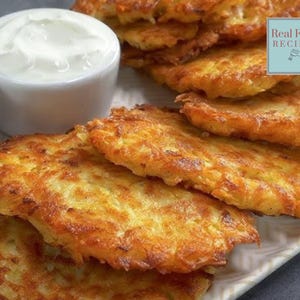 May include: A close-up of a plate of golden brown potato pancakes, also known as potato latkes. The pancakes are stacked on top of each other and are surrounded by a white plate. There are two bowls of condiments in the background, one with white sauce and one with red sauce.
