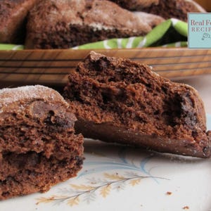 Op de afbeelding: Close-up van twee chocoladescones met chocolade chips, een is doorgebroken om de binnenkant te laten zien. De scones zijn bestoven met poedersuiker en liggen op een wit bord met een bloemenpatroon. De tekst "Real Food Recipes" is zichtbaar op de achtergrond.