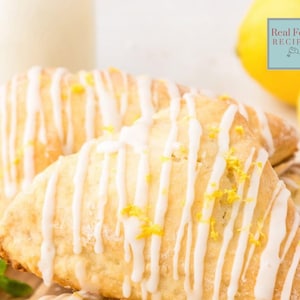 May include: Close-up of lemon scones with a white glaze and lemon zest. The scones are arranged on a wooden surface with a glass of milk and lemons in the background. The text "Real Food Recipes" is visible in the top right corner of the image.