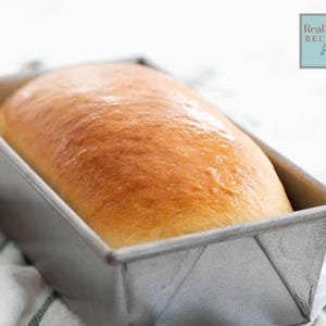 Könnte beinhalten: Ein frisch gebackenes Brot in einer rechteckigen Metallbackform. Das Brot hat eine goldbraune Kruste und eine leicht abgerundete Oberseite. Die Form steht auf einem weiß-grün gestreiften Tuch. Das Bild enthält den Text "Real Food Recipes".