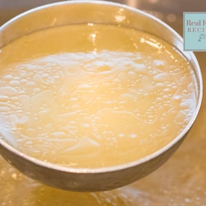May include: A close-up of a ladle filled with clear, golden broth. The broth has a slight sheen and small bubbles on the surface. The ladle is made of stainless steel and has a long handle.