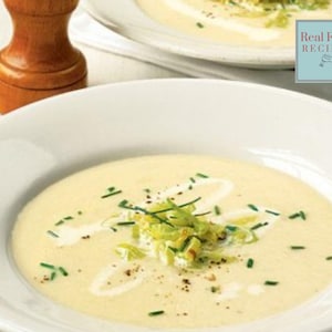 May include: A white bowl filled with creamy leek soup topped with chopped chives and a swirl of cream. The soup is garnished with sauteed leeks and black pepper.