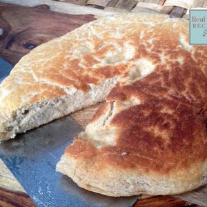 Könnte beinhalten: Ein rundes, goldbraunes Brot mit einer leicht unebenen, strukturierten Kruste. Ein großes Stück wurde herausgeschnitten und gibt den Blick auf das helle, luftige Innere frei. Ein kleines quadratisches Logo zeigt Real Food RECIPES.