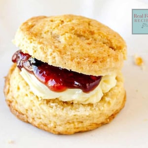 Op de afbeelding: Een goudbruine scone, doorgesneden en gevuld met een laag witte room en een royale topping van rode jam. De scone ligt op een wit bord. Het logo "Real Food Recipes" is zichtbaar.