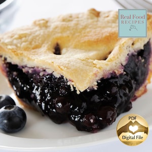 Puede incluir: Una porción de tarta de arándanos con corteza dorada y relleno de fruta de color púrpura oscuro en un plato blanco, acompañada de dos arándanos frescos. La imagen incluye una etiqueta azul con el texto Real Food RECIPES y un sello dorado.