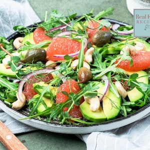 May include: A salad on a dark plate featuring sliced avocado, pink grapefruit segments, red onion rings, whole cashews, capers, and fresh arugula greens. A small square label in the top right corner displays the text Real Food RECIPES.