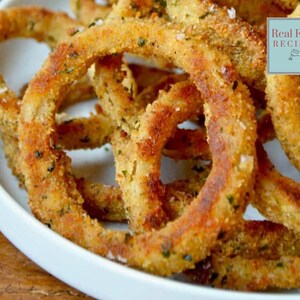May include: A pile of golden-brown breaded onion rings on a white plate, seasoned with green herbs and coarse salt. A light blue square label in the upper right corner features the text Real Food RECIPES with a small whisk icon below it.