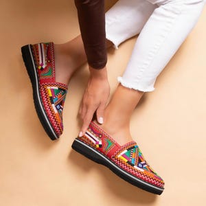 May include: Red slip-on shoes with vibrant geometric patterns. The shoes feature a black sole with a white stripe and are made of a textured material. The design incorporates yellow, green, blue, and white.
