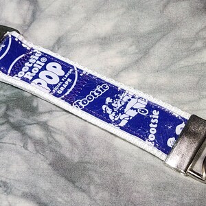 May include: A white and blue key chain with a silver clasp and ring. The key chain features a Tootsie Roll Pop wrapper with the text "Tootsie Roll Pop" and "Original Flavor Grape".