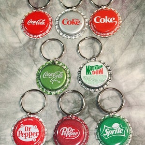 Puede incluir: Una colección de llaveros hechos con tapas de botellas de refrescos. Los llaveros presentan logotipos de Coca-Cola, Coke, Dr. Pepper, Sprite y Mountain Dew. Cada tapa está unida a un llavero plateado.