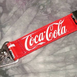 May include: Red Coca-Cola keychain with white logo text. The keychain features a silver metal clasp and key ring. The background is a gray and white tie-dye pattern.