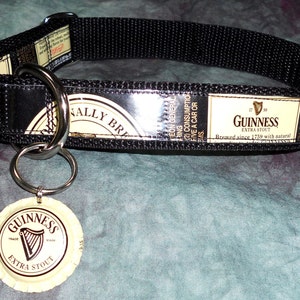 May include: Black dog collar featuring Guinness Extra Stout branding. The collar has a black nylon strap with a metal buckle and a metal ring for attaching a leash. A Guinness bottle cap is attached to the collar.
