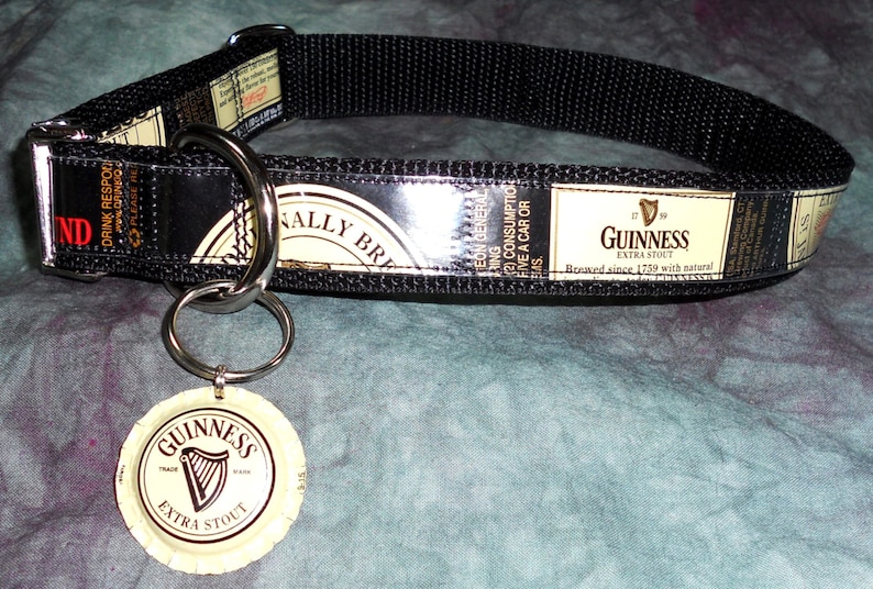 May include: Black dog collar featuring Guinness Extra Stout branding. The collar has a black nylon strap with a metal buckle and a metal ring for attaching a leash. A Guinness bottle cap is attached to the collar.