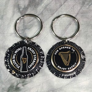 Pu&ograve; includere: Due portachiavi a forma di tappo di bottiglia. Uno presenta una grafica di bottiglia con la scritta "FROM THE BOTTLE ENJOY STRAIGHT". L'altro ha una grafica di arpa con la scritta "USE OPENER TO ENJOY SAFELY". Entrambi hanno portachiavi argentati.