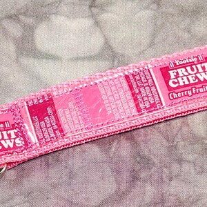 May include: Pink keychain with a silver clip and ring. The keychain is made from a pink fabric with the words "Fruit Chews Cherry Fruit Roll" printed on it.