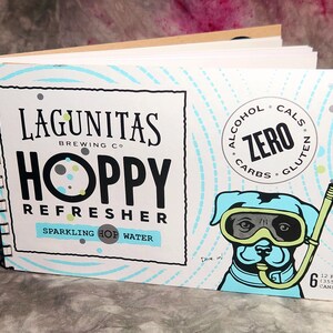 May include: A six-pack of Lagunitas Hoppy Refresher Sparkling Hop Water. The cans are blue and white with a cartoon dog wearing goggles and a snorkel. The text on the can reads "Alcohol Zero, Cals Zero, Carbs Zero, Gluten Zero".