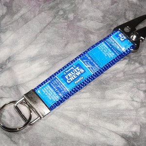 May include: Blue and silver key chain with a silver key ring and a silver clip. The key chain is made of fabric and has a blue and white wrapper with the text "Totsie II Fruit Chews Vanilla" printed on it.