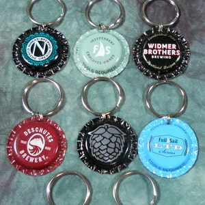 Puede incluir: Diez llaveros diferentes con forma de tapón de botella con varios logotipos de cervecerías. Los llaveros están sobre una superficie azul verdosa. Los logotipos incluyen Hub, Widmer Brothers, Deschutes, Full Sail, Rogue y Morimoto.
