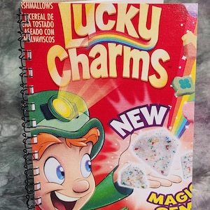 Puede incluir: Una caja de cereal Lucky Charms roja y amarilla con un duende sosteniendo una pieza de cereal blanca en forma de diamante. La caja dice "Nuevas Gemas Mágicas" y tiene un arcoíris.