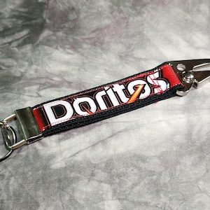May include: A fabric keychain featuring the Doritos logo in white and orange text on a red and black background. The strap is secured with silver-toned metal hardware, including a flat split ring on one end and a spring-loaded lobster clasp on the other.