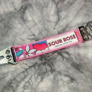 May include: A pink keychain strap with a floral and heart pattern, and the words "SOUR ROSE FRUITED SOUR ALE". The strap has a silver metal clasp and key ring.
