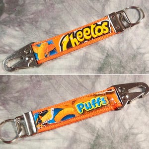 May include: Two orange keychains with silver metal clasps. One keychain features the word "Cheetos" in a white and blue font. The other keychain features the word "Puffs" in a white and blue font.