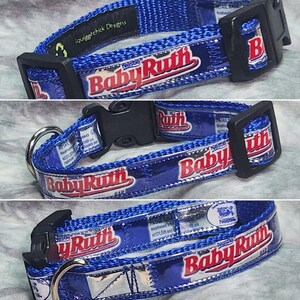 May include: A blue pet collar featuring the Baby Ruth candy bar logo in red and white text. The collar is constructed from woven nylon webbing with a black plastic side-release buckle and a silver metal D-ring for leash attachment.