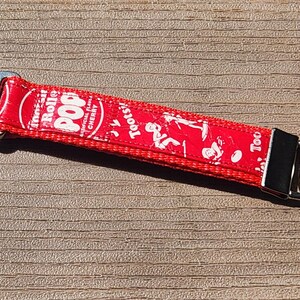 May include: A red fabric keychain featuring a repeating white graphic print of the Tootsie Roll Pop logo and cherry candy imagery. The strap is secured with a silver-toned metal lobster clasp on one end and a circular split ring on the other.