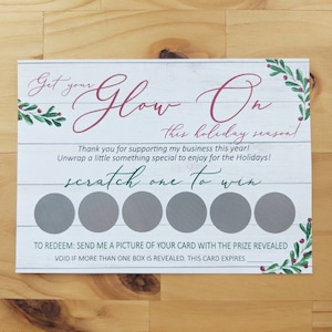 May include: A white card with a woodgrain background and red and green holly sprigs. The text "Get your Glow On this holiday season!" is written in pink script. The card has six gray scratch-off circles with the text "scratch one to win" below. The text "TO REDEEM: SEND ME A PICTURE OF YOUR CARD WITH THE PRIZE REVEALED VOID IF MORE THAN ONE BOX IS REVEALED. THIS CARD EXPIRES" is printed at the bottom.