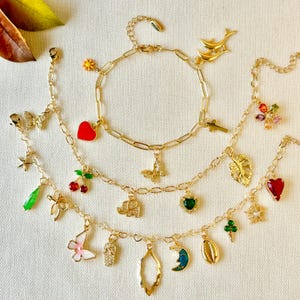 May include: Three gold chain bracelets with various charms, including a red heart, a green leaf, a butterfly, a star, a cherry, a green heart, a seashell, a crescent moon, a pineapple, a cross, a dolphin, a monstera leaf, an elephant, and a starfish.