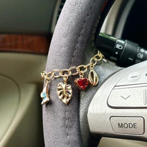 Peut inclure: Un bracelet de charme doré avec divers pendentifs, dont un papillon, une feuille, un cœur et un coquillage. Le bracelet est attaché à un volant. Le pendentif en forme de cœur est rouge.