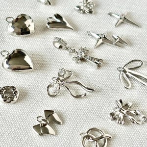 May include: A collection of silver charms and pendants, including hearts, bows, crosses, and other decorative shapes. The charms are displayed on a white, textured surface, showcasing their intricate designs and reflective surfaces.