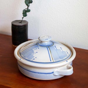 May include: A ceramic lidded casserole dish with a blue and white design. The dish has a round shape with a handle and a matching lid. A small black vase with a sprig of greenery is behind the dish.