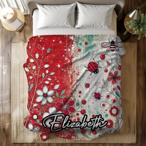 May include: A red and white floral blanket with the name "Elizabeth" in a cursive font. The blanket features a design of flowers, ladybugs, and a bee. The blanket is on a bed with white pillows.
