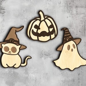 Puede incluir: Tres decoraciones de Halloween de madera: una calabaza con una cara tallada, un fantasma con un sombrero puntiagudo y un gato con un sombrero de bruja.