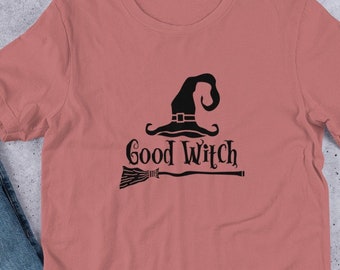 Può includere: T-shirt rosa polveroso a maniche corte con una grafica nera di un cappello da strega, una scopa e le parole "Good Witch". La maglietta è realizzata in un materiale morbido ed è adatta per l'abbigliamento casual. Il design è centrato.